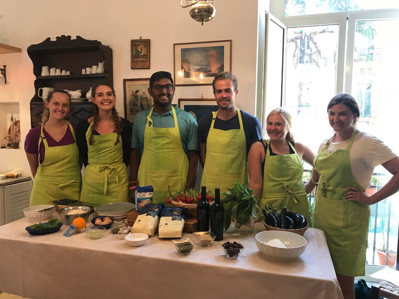 Six students pose in front of wine and cooking supplies before they begin preparing an Italian meal at a cooking class in Napoli.