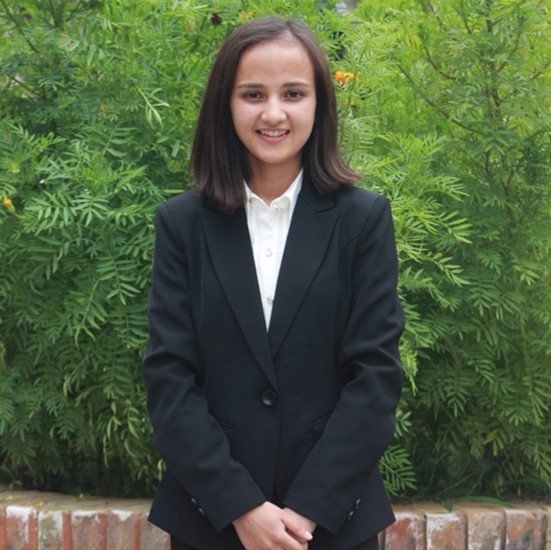 A smiling color photo of Sirjana Pun wearing a black suit in front of a leafy background.