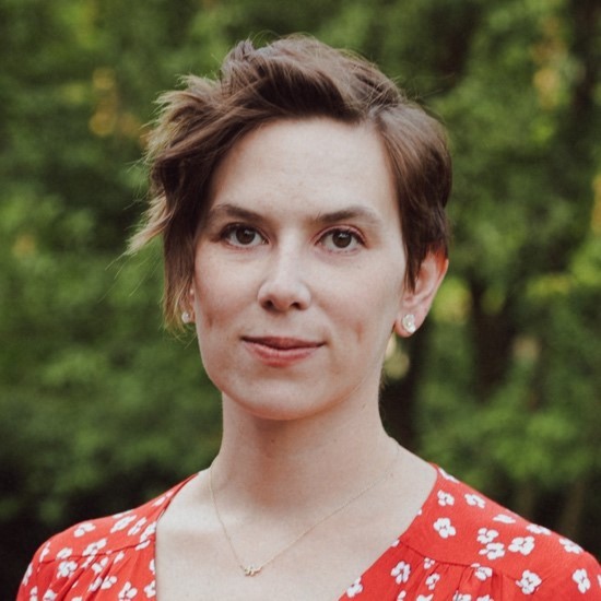 A smiling color photo of Corinne Jorgenson wearing a red floral dress in front of a leafy background.