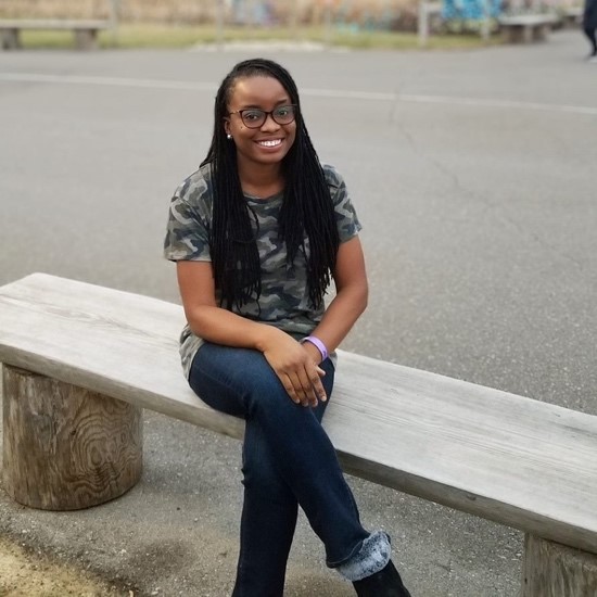 A smiling color photo of Shammah Omololu sitting on a bench wearing a camo shirt and jeans.