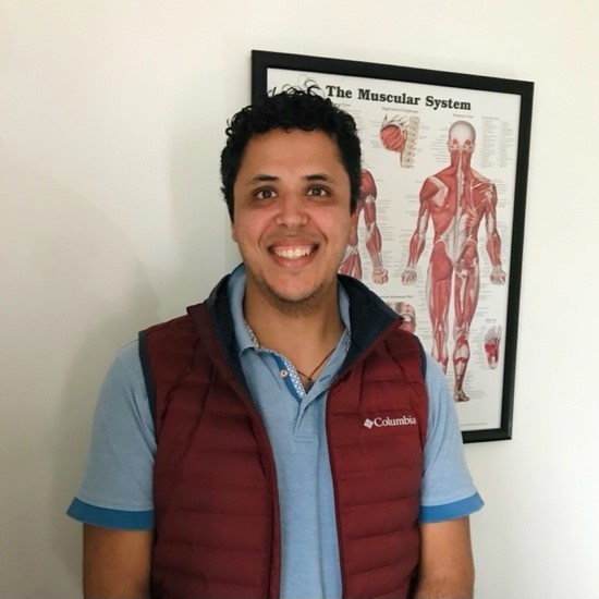A color photo of Tarcisio Moreira smiling, wearing a red vest and a blue shirt and standing in front of a poster that says "the muscular system."