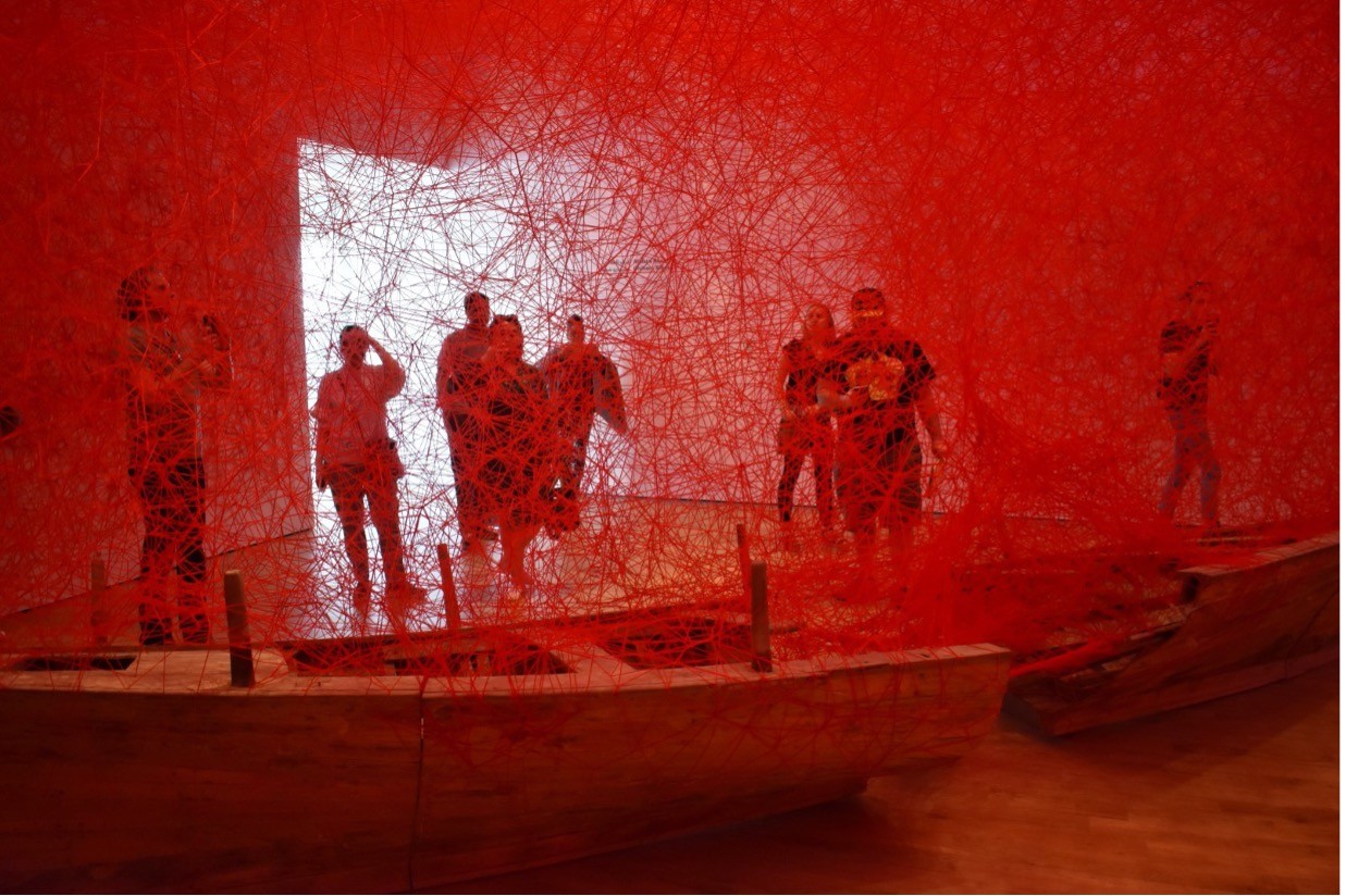 A room is filled with red webbing over a long boat that is split in half on the floor. A group of people stand behind admiring the webbing-and-boat artwork with a light open doorway in the background.