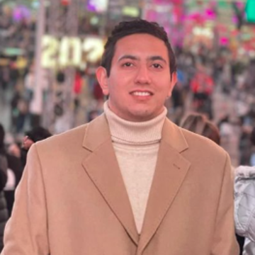 A smiling photo of Manish Raj Aryal with dark hair, wearing a beige turtule neck shirt under a light brown jacket, with a blurry and colorful crowd in the background.