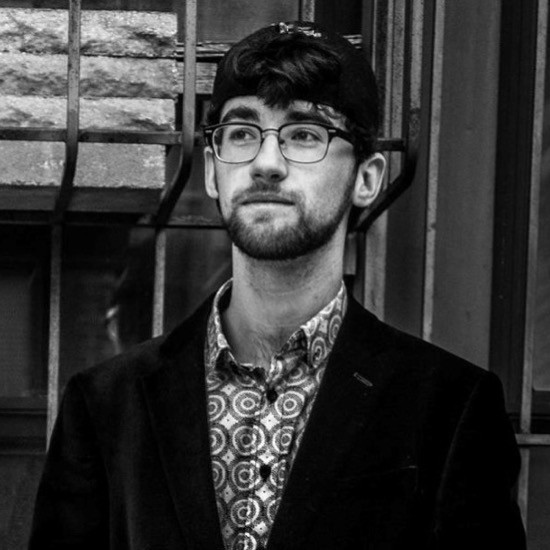 A smiling, black-and-white photo of Kieran McAuliffe wearing a bullseye-patterend, collared shirt under a dark jacket, glasses, and baseball cap.